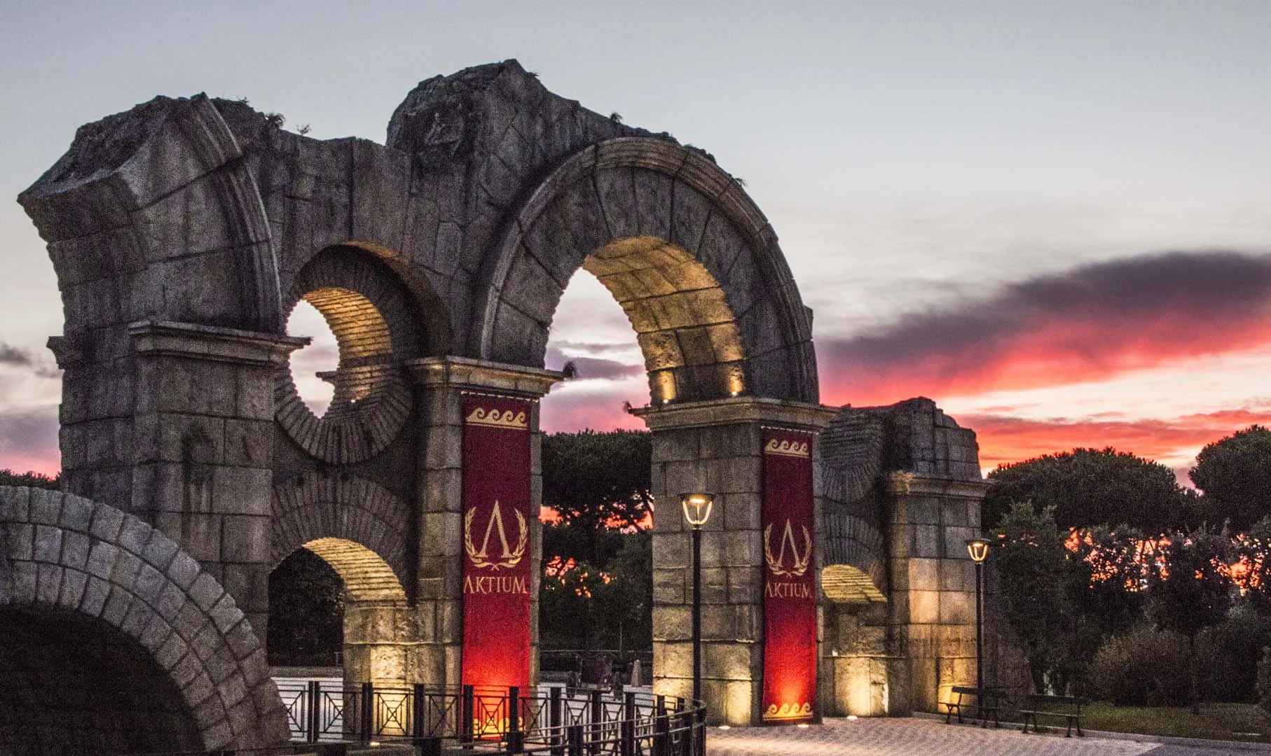 The triumphal arch of the Roma area at Cinecittà World at sunset, red-violet sky above the sculpted faux-stone arch
