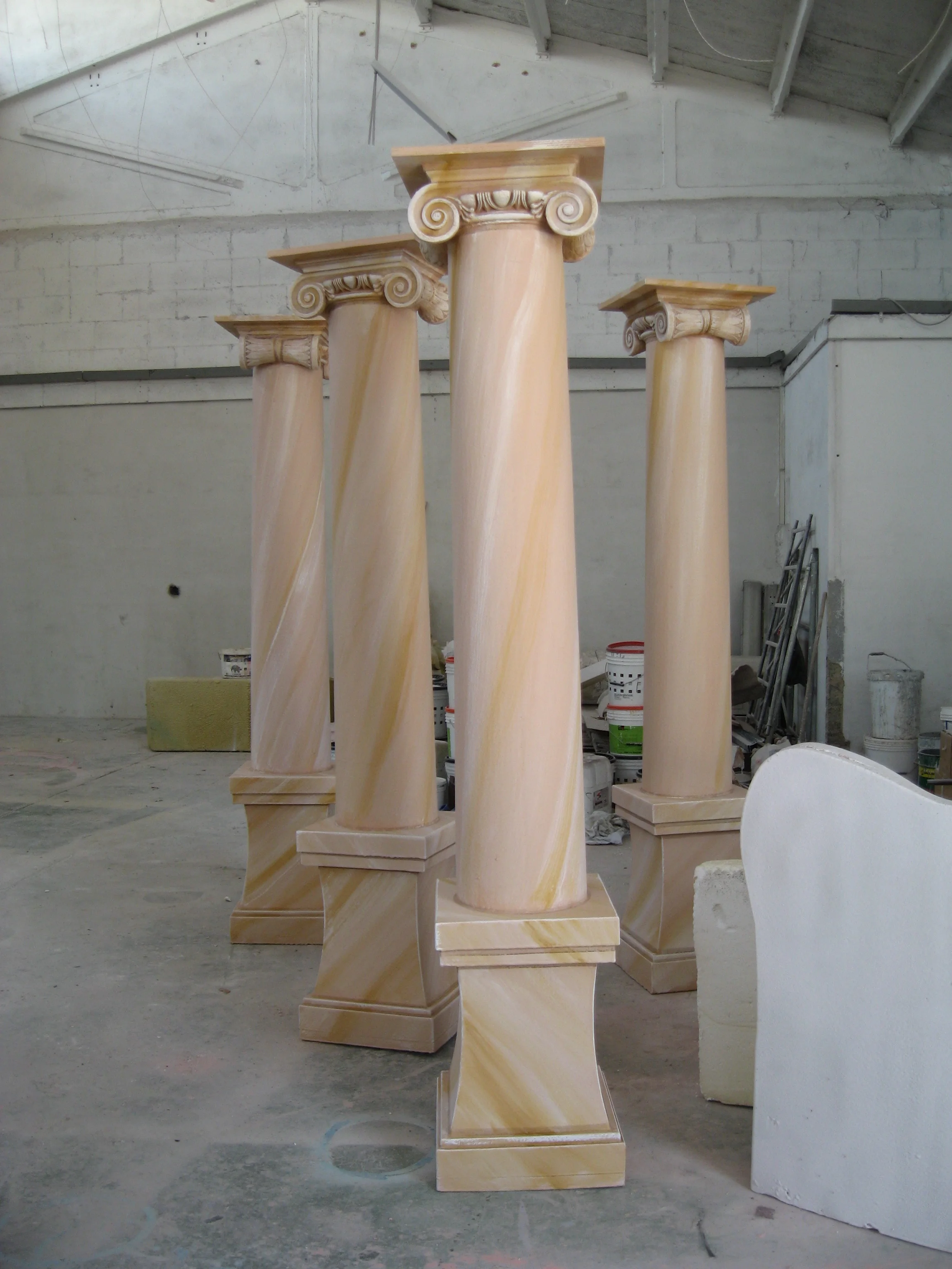 Four scenographic Ionic columns for the Cannes Film Festival in the Creation Art workshop: about three metres tall, shafts in faux onyx marbled white-and-yellow, Ionic capitals with volutes, bell-shaped bases, seen in a row waiting for transport