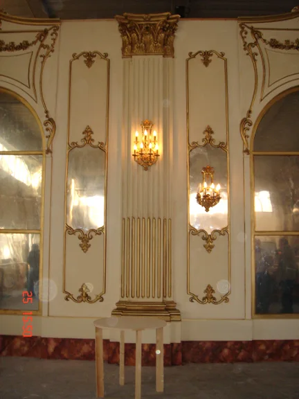 Side panel of the Sissi Hall: white wall with three baroque gilded frames in rocaille and acanthus leaves, a hanging eight-arm gold candelabrum above, two smaller gold candelabra on either side, in the foreground a temporary worksite table and chair