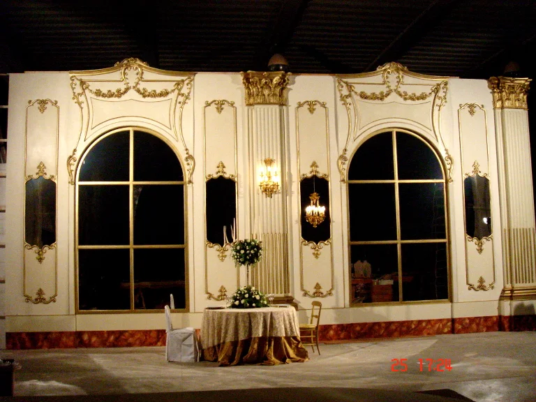 Full view of the Princess Sissi Hall rebuilt for a wedding in Dubai: white walls with baroque golden stuccoes, two large arched windows with gilded glass framing, Corinthian columns with gilded capitals on the sides, hanging gold candelabra, in the foreground a round wedding table with white floral decoration and a side chair