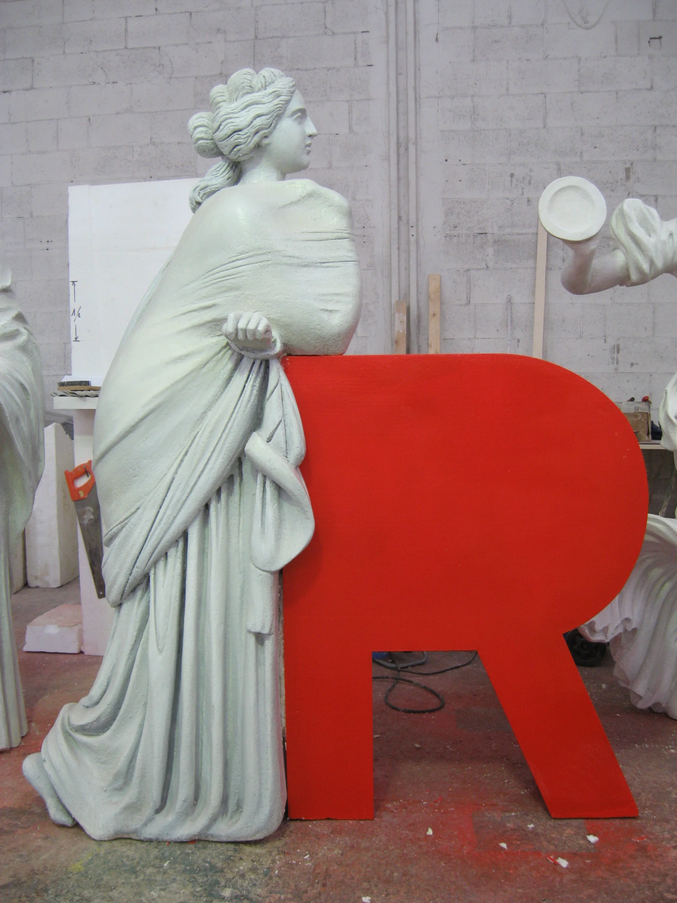 Classical statue for Galleria Sordi during construction: three-quarter-view draped female figure, sculpted polystyrene patinated in light grey, a monumental red R letter behind her, in the background the industrial hangar of the Creation Art workshop