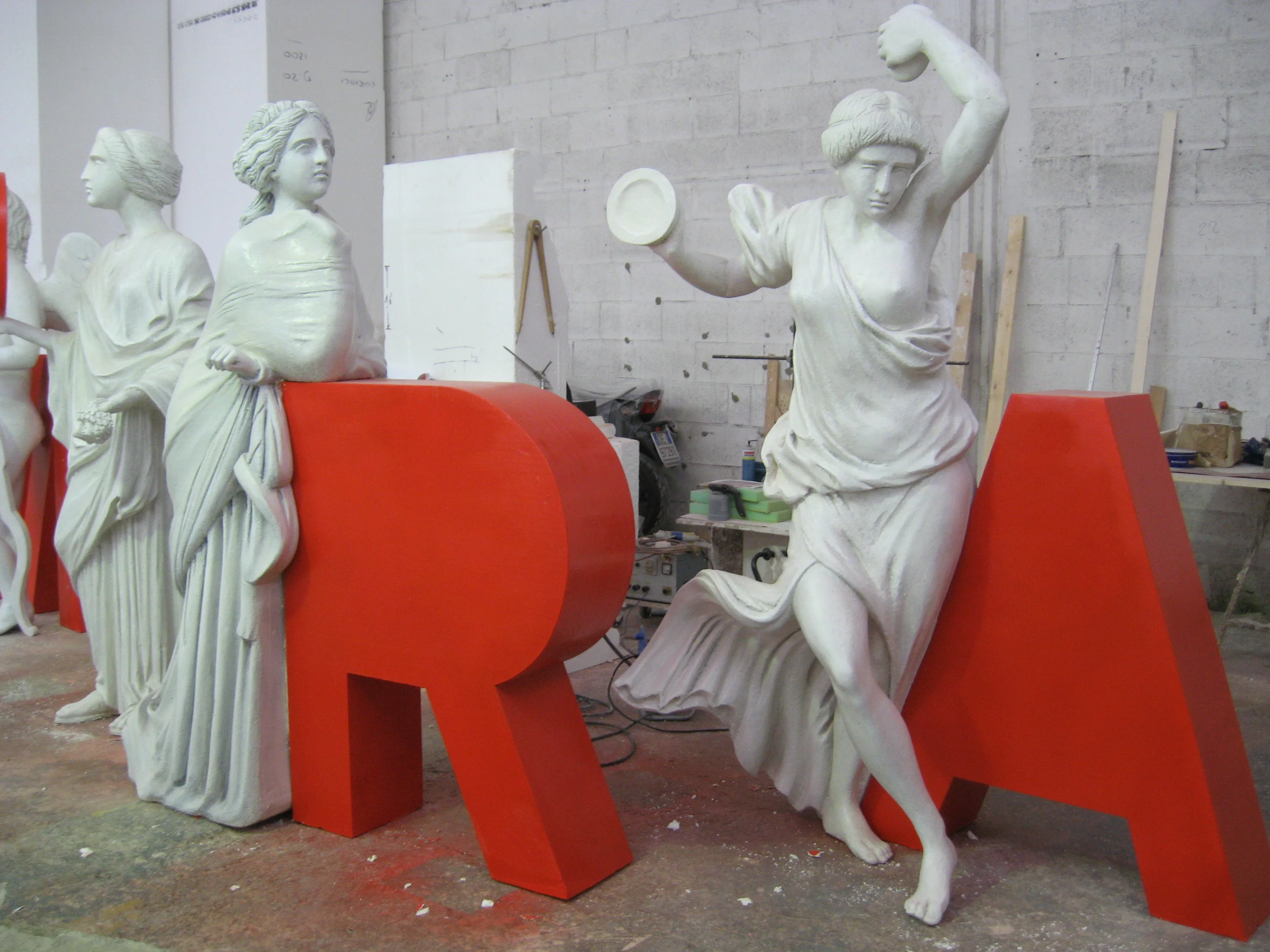 Installation for the Galleria Alberto Sordi: three white classical statues sculpted in polystyrene and fibreglass, one with a flat heart raised, two in neoclassical poses, set against bright red R and A letters in faux stone two metres tall, viewed in the workshop yard before delivery
