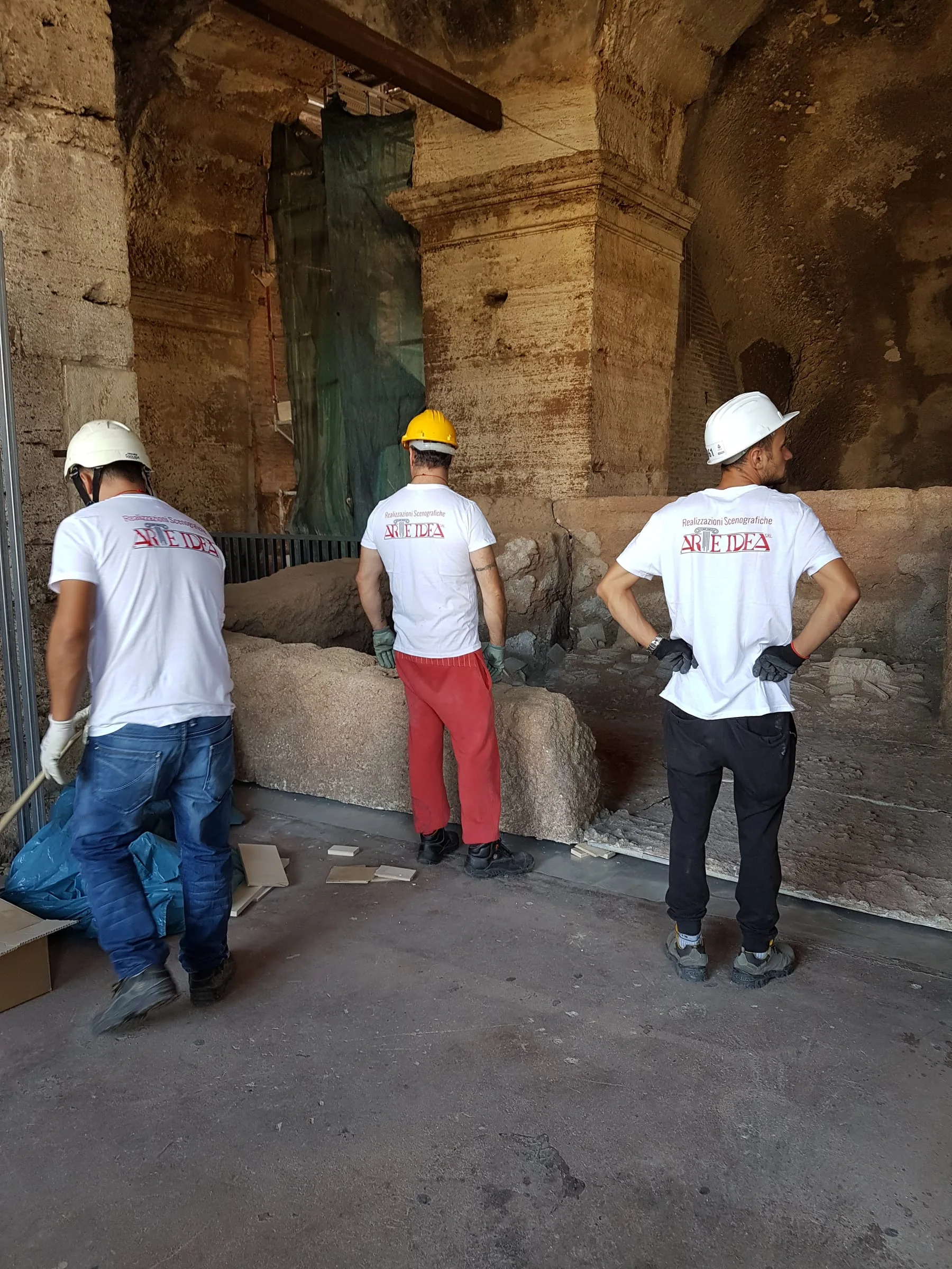 Tre operai di Creation Art con maglietta bianca con logo bordeaux installano la Sala dell'Archivio dentro gli spazi espositivi del Colosseo: scaricano un blocco di muratura e lo posizionano