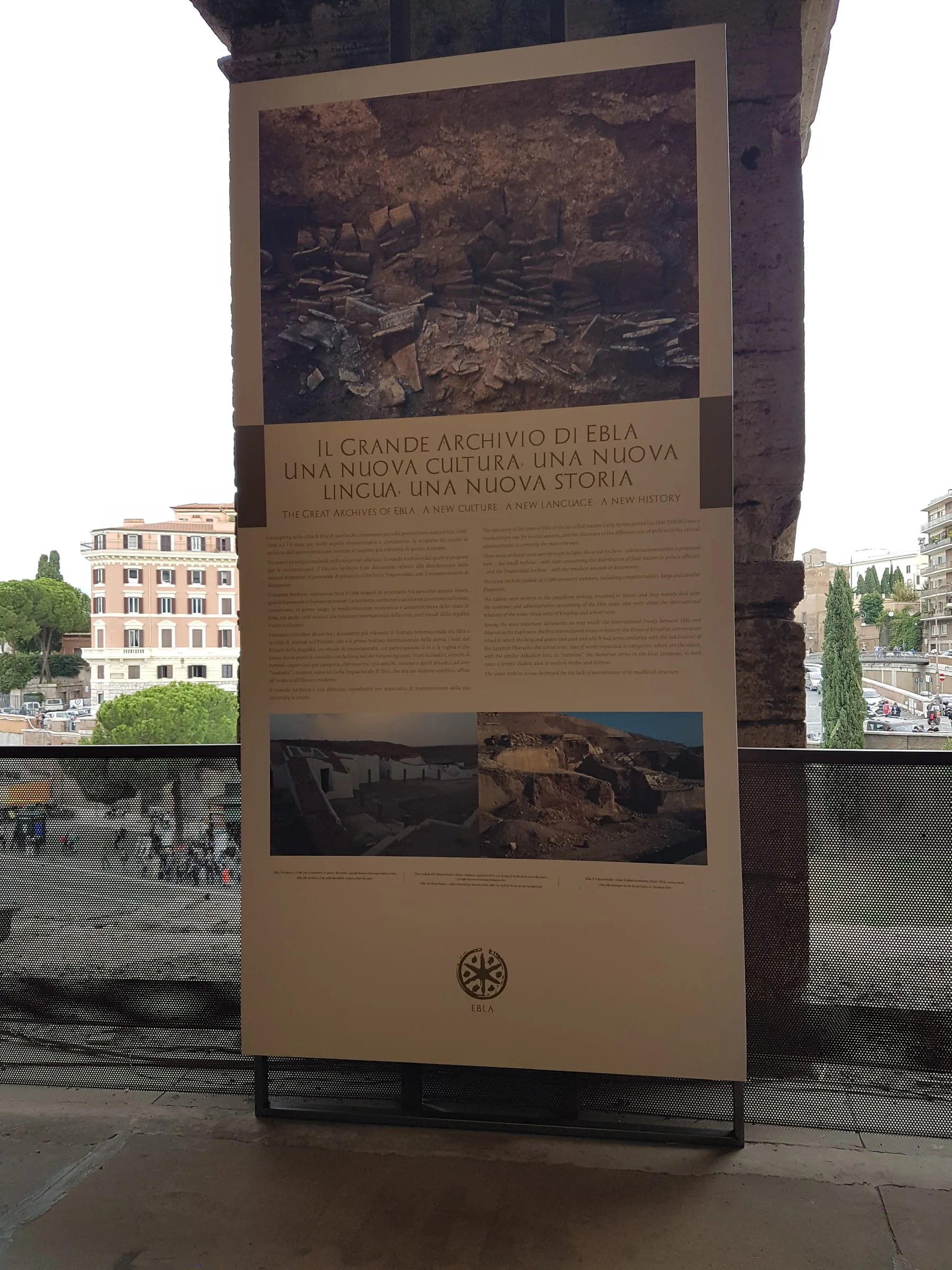 Pannello esplicativo della mostra 'Rinascere dalle distruzioni' davanti al Colosseo: titolo 'Il grande archivio di Ebla — una nuova cultura, una nuova lingua, una nuova storia', logo della mostra, foto di scavo