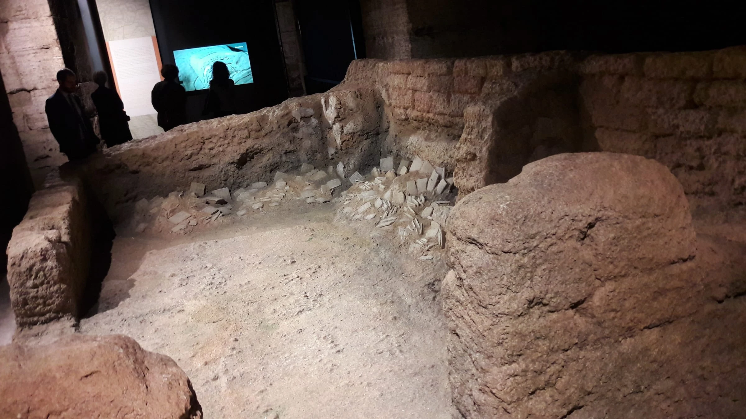Vista grandangolare della Sala dell'Archivio di Ebla installata al Colosseo: la muratura del Palazzo Reale G ricostruita 1:1, le tavolette cuneiformi cadute sul pavimento, alcuni visitatori in primo piano, monitor sullo sfondo che proietta video archeologico