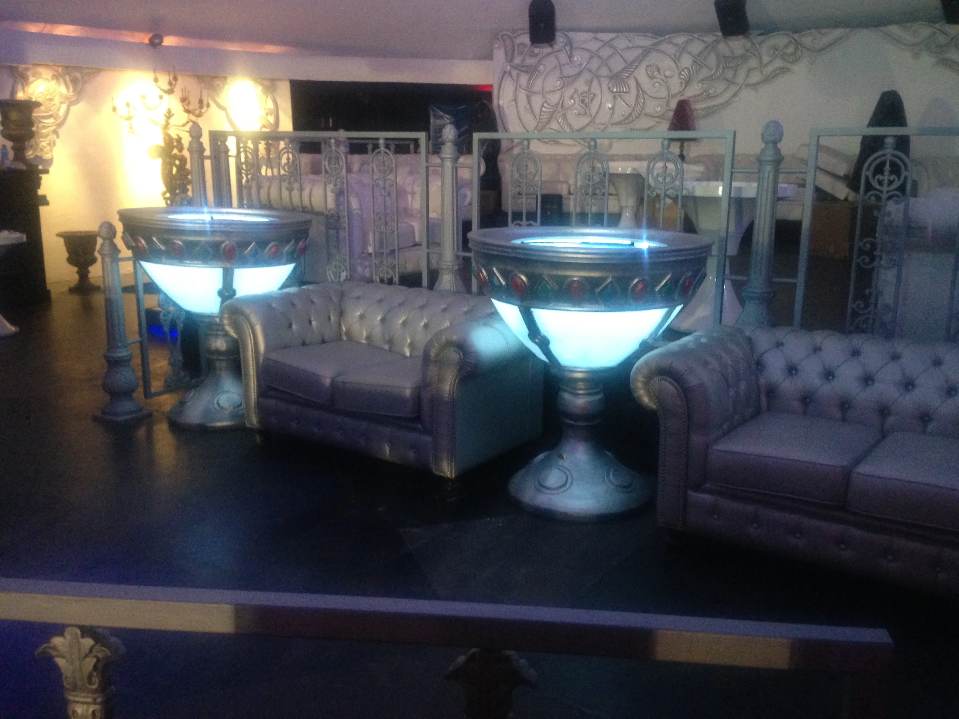 One of the monumental backlit goblets of Art Café seen from the front: an ancient-chalice shape around two metres tall, a column base decorated in relief, embossed silver metallic rims, a luminous central section in opalescent plexiglass that diffuses a cool blue-violet light, metallic-leather Chesterfield sofas visible around it