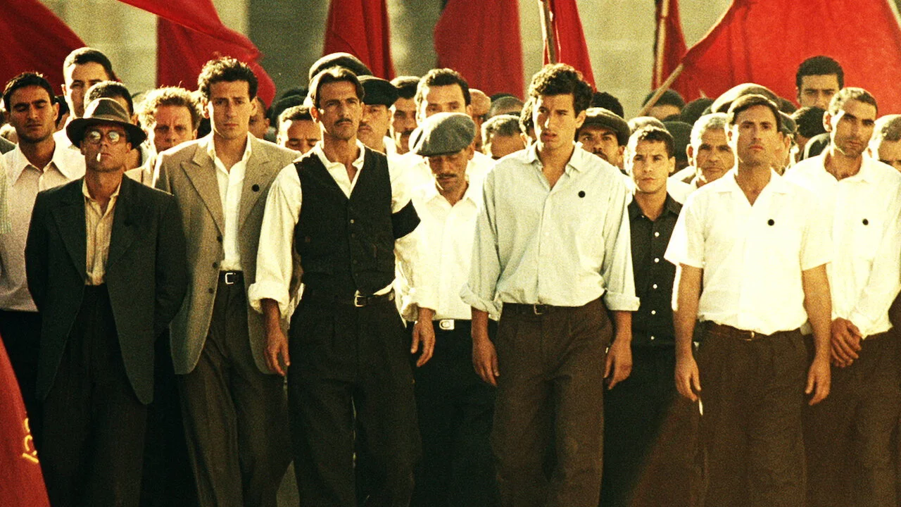 Political procession with red flags of the Italian Communist Party — men in flat caps in the foreground, a mass scene shot on the Tunisian set.