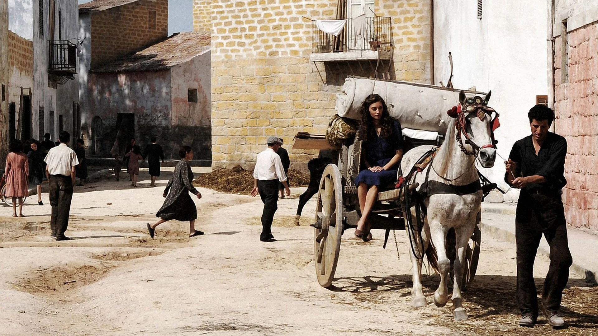 A side street in Bagheria: mustard and pink stone houses, balconies, a white horse-drawn carriage, extras in 1930s costume.