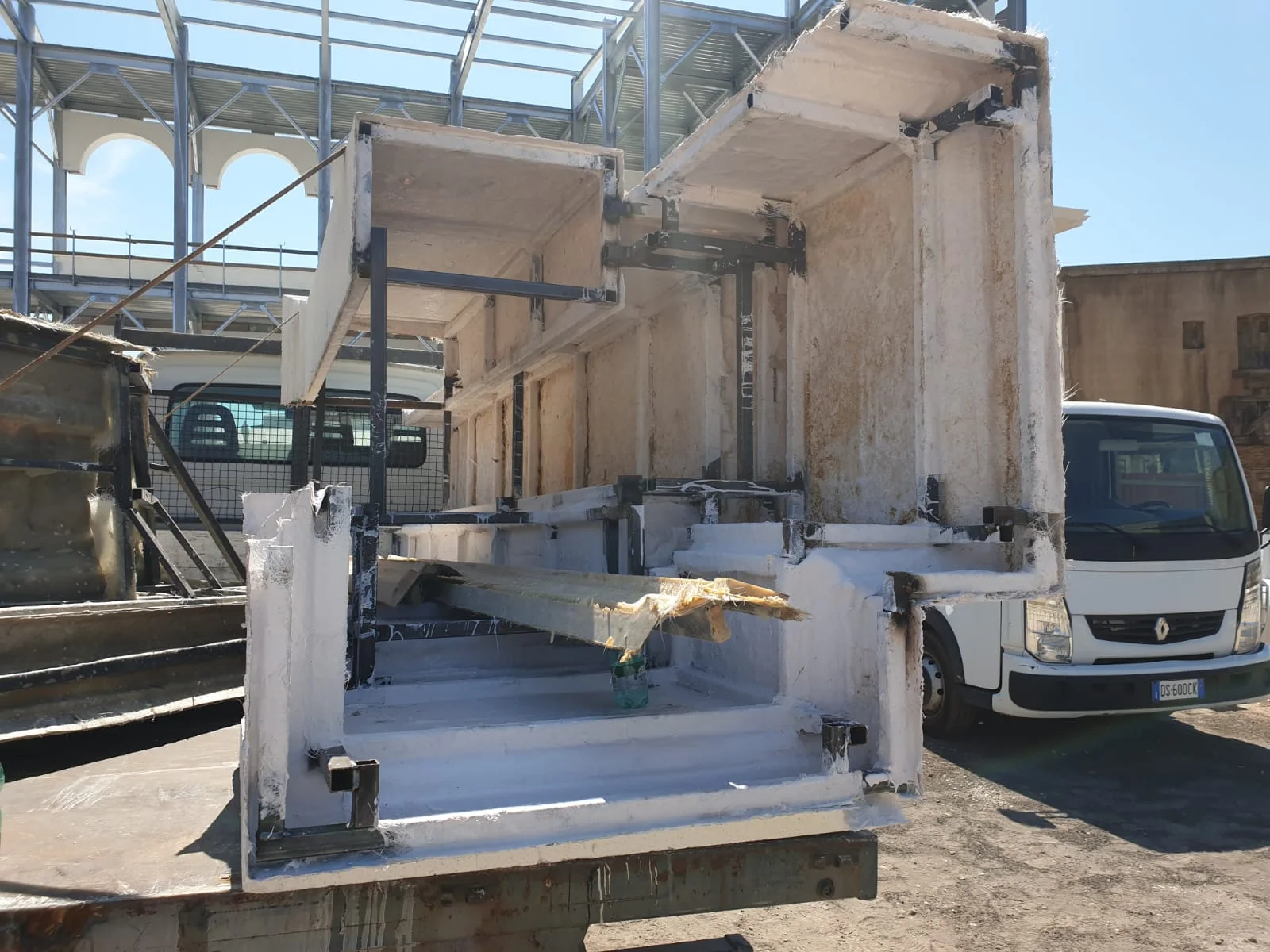 Dettaglio finale del cantiere della Basilica Giulia: pannello di vetroresina ravvicinato con incassi che simulano la mensola dentellata di un architrave classico, finitura ancora grezza pre-pittura, base posata sul terreno polveroso accanto a un piccolo segnale di cantiere
