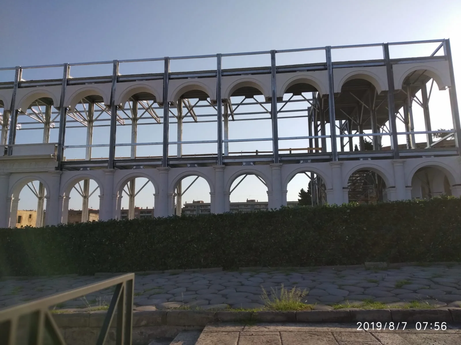 Vista del cantiere: la struttura metallica della Basilica Giulia vista frontalmente, archi del piano terra completati con pilastri in finta pietra bianca, archi del piano superiore ancora aperti come scheletro, dietro il basolato della pavimentazione del Foro romano già posato