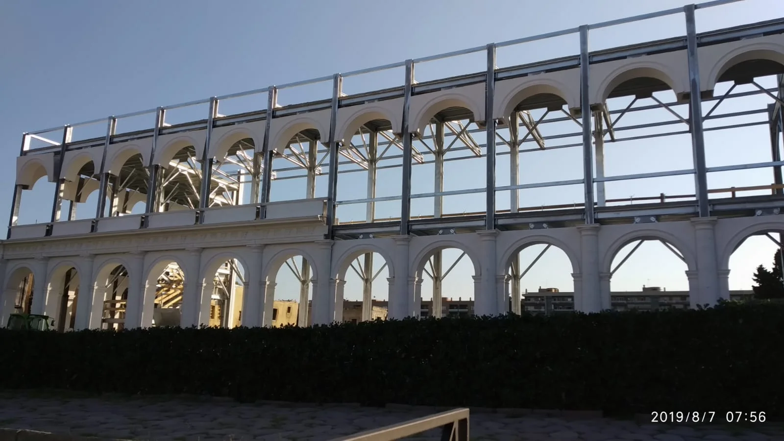Cantiere della Basilica Giulia il 7 agosto 2019, vista in controluce: struttura metallica tubolare già montata in alto, primo ordine di archi a tutto sesto in finta pietra bianca al piano terra completato, scheletro metallico del secondo piano ancora vuoto, fondo del piazzale con i palazzi residenziali visibili attraverso gli archi, cielo azzurro mattutino