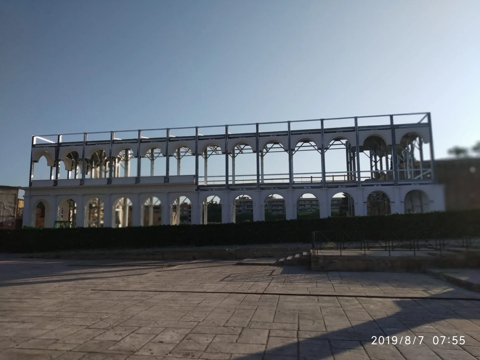 Vista panoramica della Basilica Giulia in cantiere all'alba del 7 agosto 2019: edificio a due piani con primo piano d'archi in finta pietra bianca quasi completati, secondo piano scheletro metallico visibile in tutta la sua estensione, pavimentazione del Foro a lastre già posata in primo piano, cespugli e palazzi residenziali in lontananza