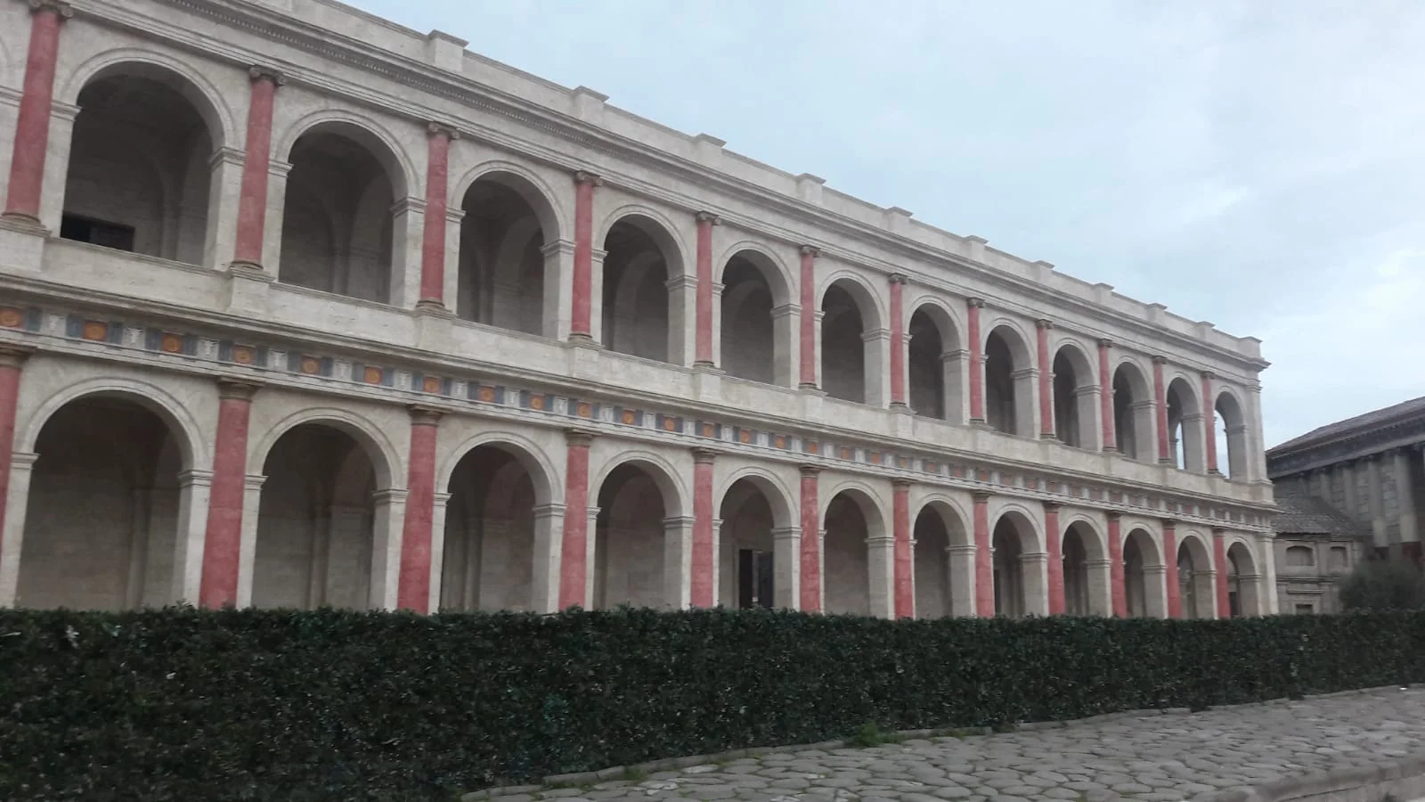 La Basilica Giulia restaurata per Domina, vista frontale: facciata a due piani con doppio ordine di archi a tutto sesto, pilastri in finto marmo rosso pompeiano alternati alla pietra travertino color avorio, fregio policromo con cerchi blu e ocra fra i due ordini, pavimentazione a lastre di pietra calpestabile, sullo sfondo a destra altri edifici del set di Roma