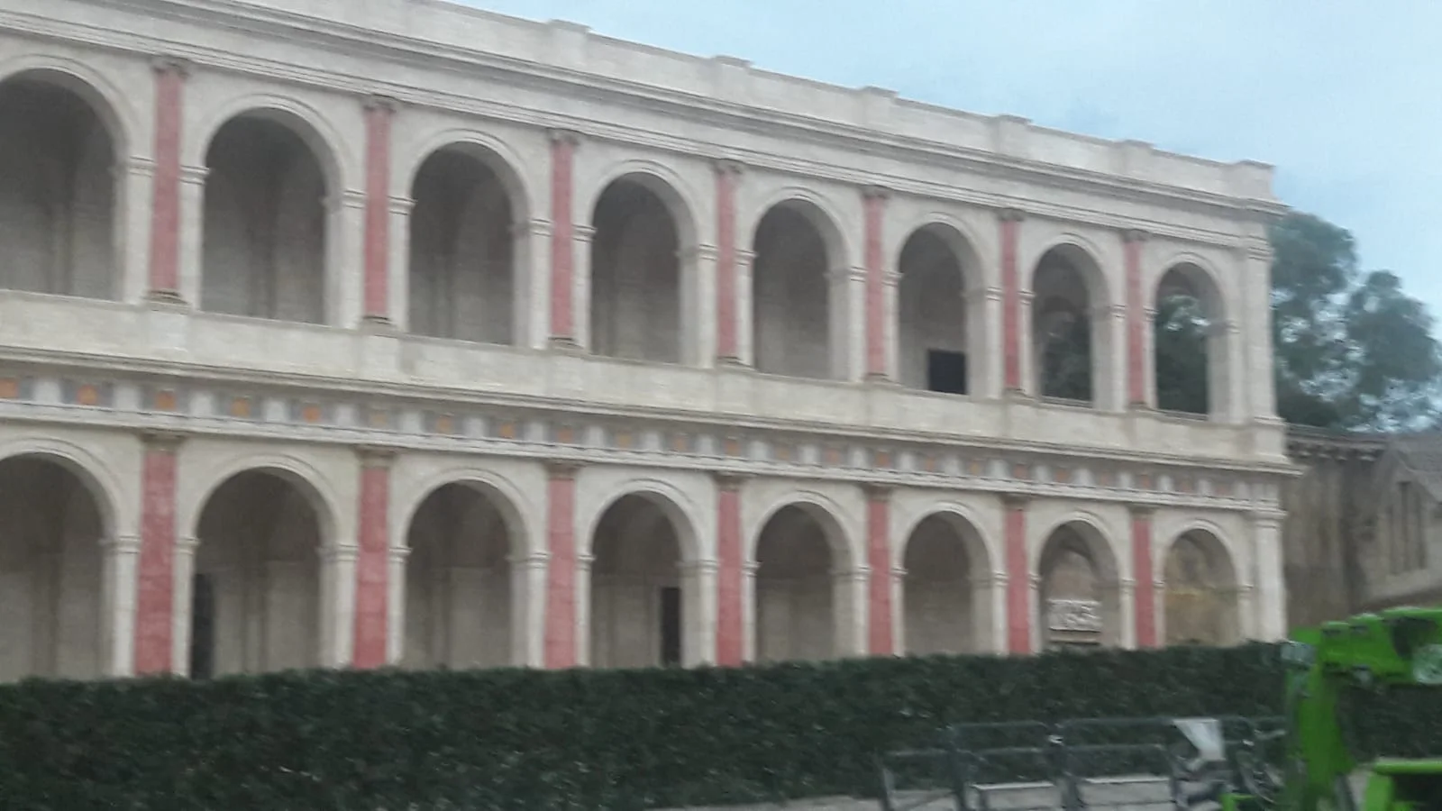 La stessa facciata vista da angolazione laterale, in luce di crepuscolo: ordinatura degli archi più ravvicinata, dettaglio delle dentellature dell'architrave, finta pietra travertino color avorio scolpita a velature, sullo sfondo gru gialla del cantiere
