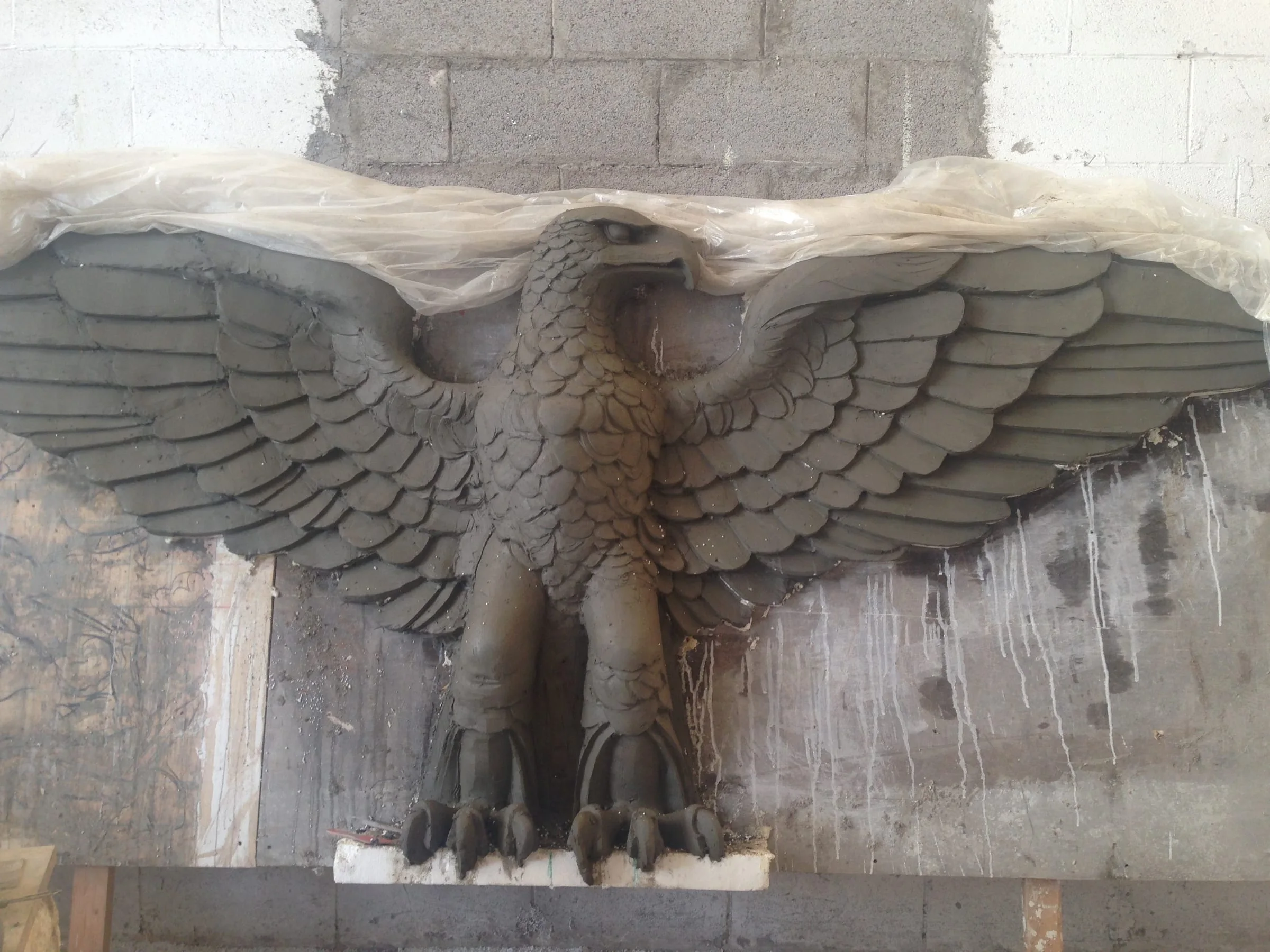 Aquila imperiale per Ben Hur scolpita in argilla creta grigia nei nostri laboratori: rapace antropomorfo con grandi ali aperte distese sui lati, becco aperto, sguardo fiero verso destra, piume delle ali modellate singolarmente; sullo sfondo parete del laboratorio in blocchi di calcestruzzo grezzo