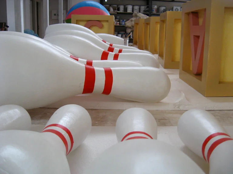 Rows of bowling pins sculpted in fibreglass: classic bowling-bottle shape, white with three red stripes near the top, laid horizontally in the workshop, alphabet cubes in yellow and pink with letters S and A visible in the background