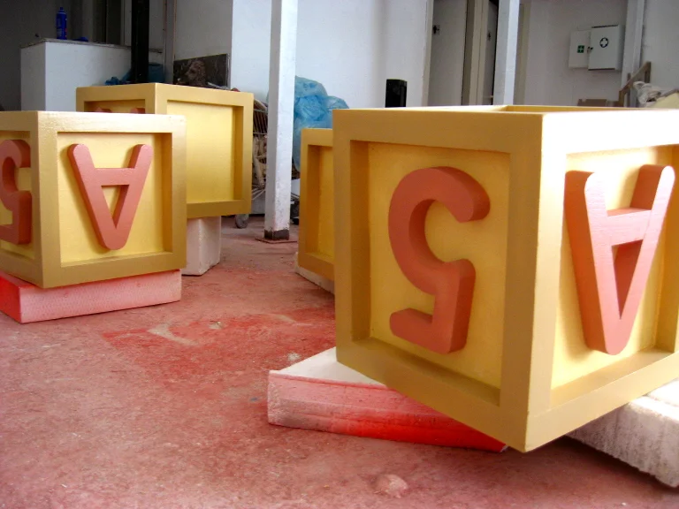 Giant alphabet cubes in yellow and pink with sculpted letters and numbers (A, S, V, 5), pink bases, lined up in the workshop yard
