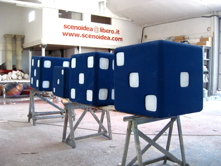 Three giant blue dice sculpted in fibreglass, each with white sculpted dot faces, around a metre per side, mounted on wooden trestles in the warehouse, with the SCENOIDEA logo visible on the wall behind