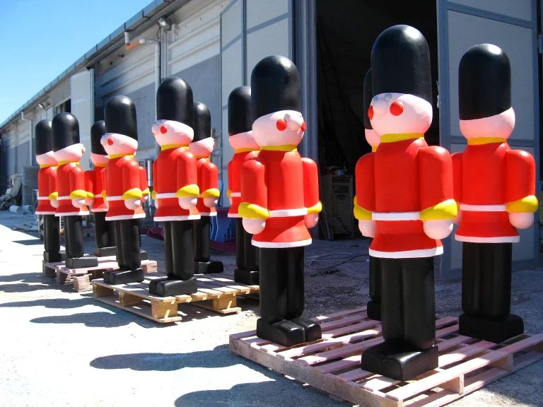 Seven toy British royal guards in the workshop yard: red cloak with yellow buttons, round pink face with red cheeks, tall black bearskin hat, all standing on wooden pallets with the industrial workshop in the background
