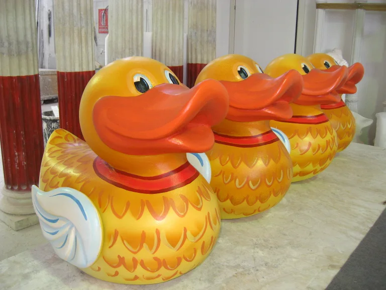 Four giant yellow toy ducks sculpted in fibreglass: golden body with sculpted feathers, wide orange beak, round black eyes, lined up like a fairground duck-shoot