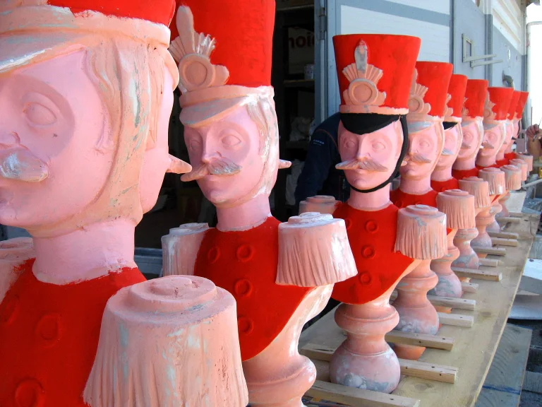 Row of nutcracker-soldier busts in finishing stage: round head with white moustache, tall red tube hat with feather and gold trim, red military uniform with buttons, lined up in the yard before the final paint coat