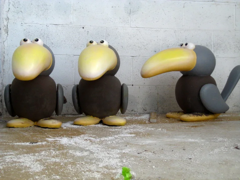Three stylised cartoon birds sculpted in fibreglass: round brown bodies with grey wings, round black eyes, yellow beaks — two with short rounded beaks on the left, one with a long toucan beak on the right, set in the workshop warehouse