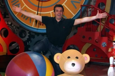Flavio Insinna with arms wide open at the centre of the set of Il Braccio e la Mente: playful pose between the yellow-and-red ball on the left, the brown teddy bear with pink heart in the middle, the British guard on the right, with the large orange retro clock and red cogwheels in the background