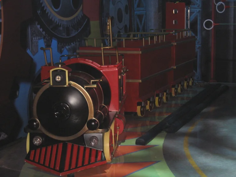 The stylised steam locomotive installed in the studio: round black nose with lamps, red cage, red flatbed wagons, with a painted backdrop of large blue and red cogwheels and gears behind