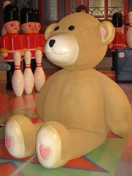 The large brown teddy bear sitting on the studio floor: round head, black eyes, pink heart under the sole of its foot; behind it the toy British guards lined up and bowling pins, floor in coloured triangles
