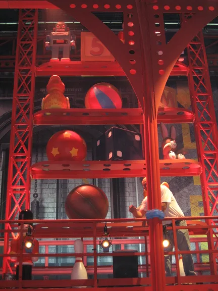 The set installed in the studio: red industrial structure with beams and rivets, two-tier shelving, on the shelves one of the blue robots, an alphabet cube, balls, a Pac-Man silhouette, a pawn shape, a basketball, bowling pins; a technician climbing a ladder on the right