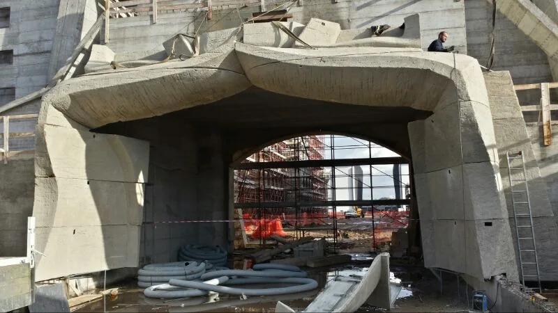 The central arch of the gate seen from the interior during construction: giant vesica piscis (almond) shape, wavy concrete structure being finished, paint cans and pipes on the ground, workers above