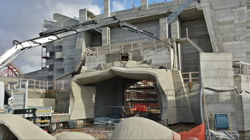Front overview of the gate under construction: central vesica piscis arch, side ribs, yellow telescopic crane on the right, construction rubble in the foreground