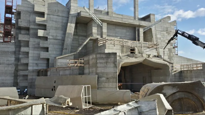 3/4 view of the gate's side façade: concrete ribs, curved wavy entrance arch, monumental staircase, construction site still active with machines in the background