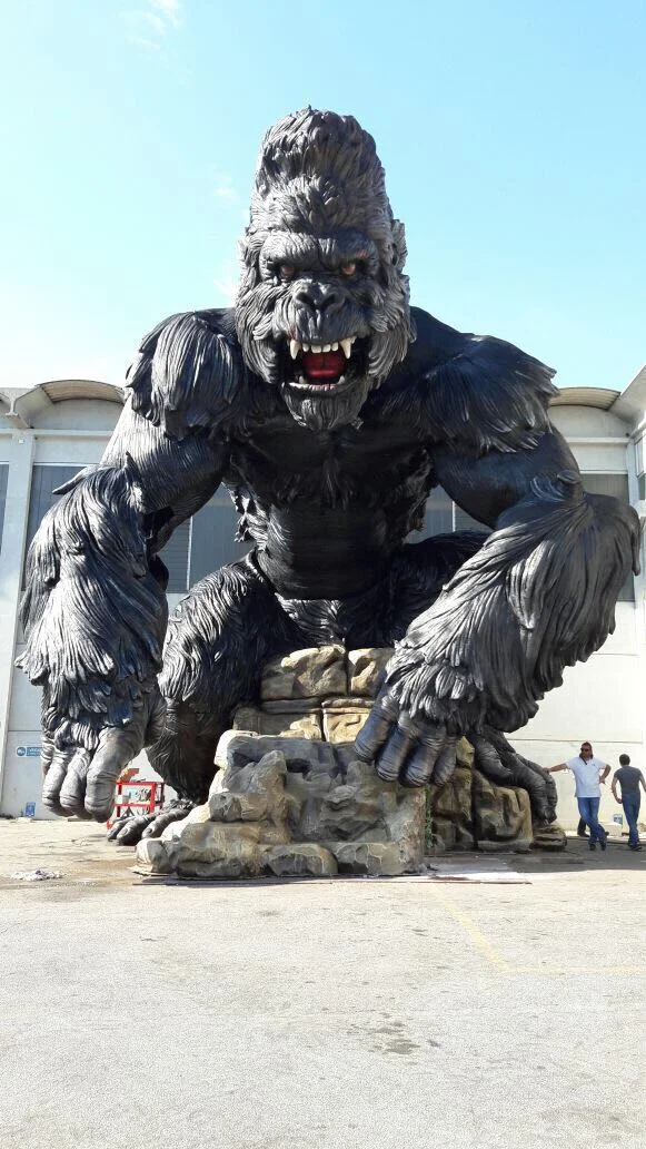 The finished giant King Kong in our workshop yard: monumental sculpture about eight metres tall in black-metallic fibreglass, profile of the gorilla seated on the fake-stone boulder, arms forward, jaws open with white teeth and red tongue, red eyes lit, fur carved in dark tufts; a person in the right foreground for scale, industrial warehouse in the background
