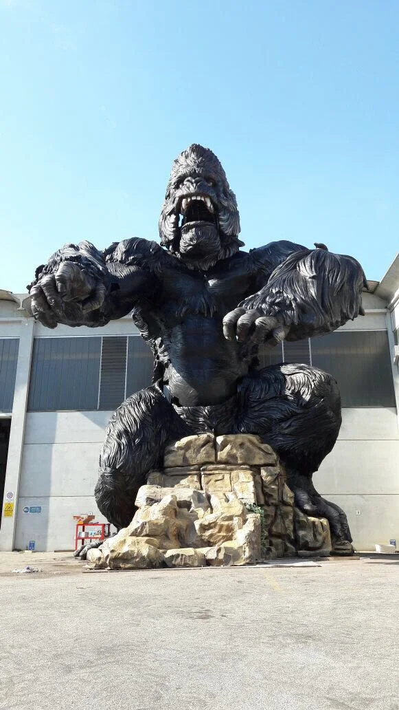 Another angle of King Kong in the yard: low-angle view emphasising the mass of the sculpture, muscular arms reaching out from the sides, wide-open jaws with teeth, fur carved in tufts across the whole body, the fake-stone base on which it sits, the Tunisian sky in the background