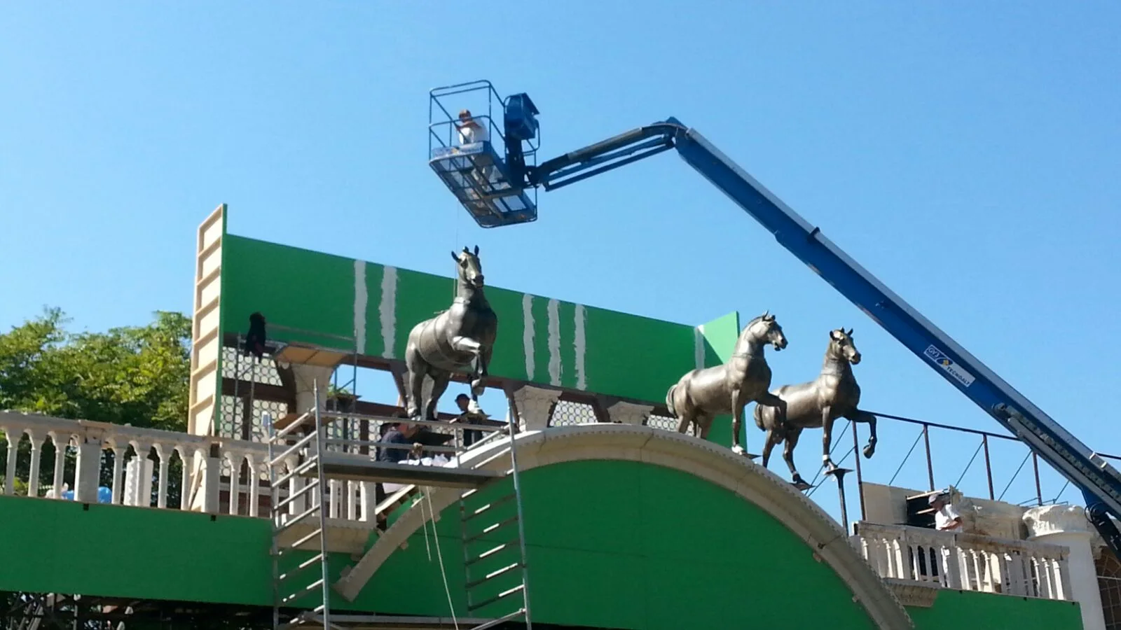 Tre quadrighe già montate sopra l'arco della Loggia dei Cavalli ricostruita: fondale verde di chroma key sullo sfondo, operai su cestello sollevatore che terminano l'installazione, struttura in finto marmo bianco