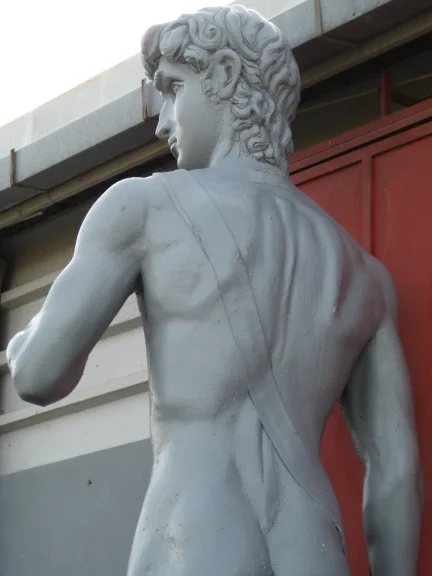 Replica of Michelangelo's David in a gray-white scenic finish: head with classical curls, heroic profile, three-quarter rear view, displayed in the workshop yard, red industrial background