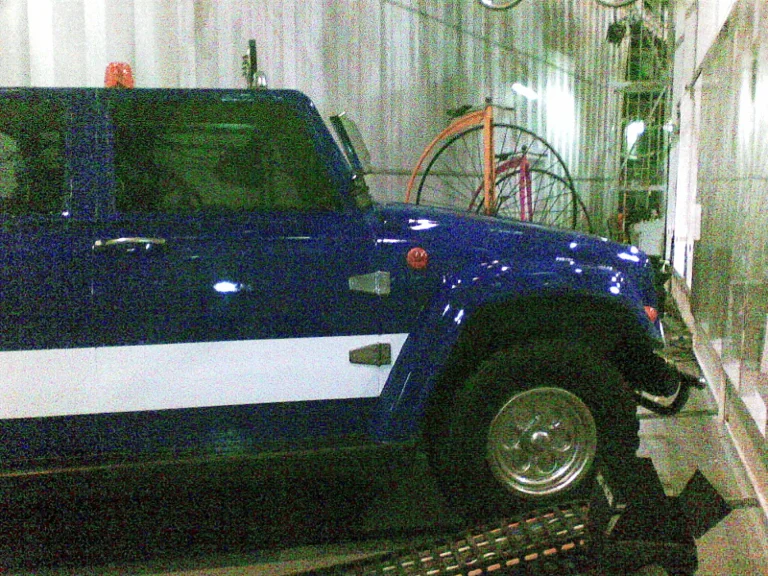 The blue car seen from the side: off-road profile with horizontal white stripes, glossy metallic blue bodywork, being worked on in the workshop behind white drapes, a red vintage bicycle in the background