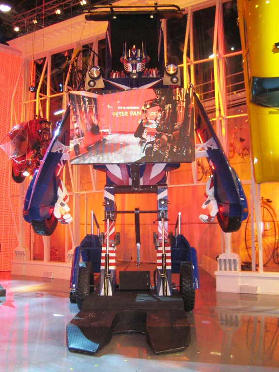The transformer robot open seen from the front in the studio: blue/white body with a British flag, red and blue side wings, Optimus head, LED screen in the chest reading PETER PAN, chassis wheels below, industrial structure in the background
