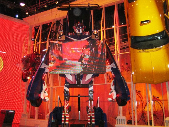 The transformer robot open in the studio: blue and white striped body with a British flag, side arms with elements of the vehicle's bodywork, Optimus Prime-style head with visor, a large LED screen in the chest showing the Peter Pan logo and the show's animation, stage lights above, yellow lattice structure in the background