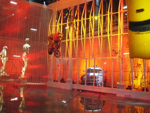 Panoramic view of the set installed in the studio: at the center the closed blue car parked, to the sides two large white classical statues (David and a hero with scepter), above the audience a red hot-air balloon and a biplane suspended, yellow industrial lattice structure against an orange background