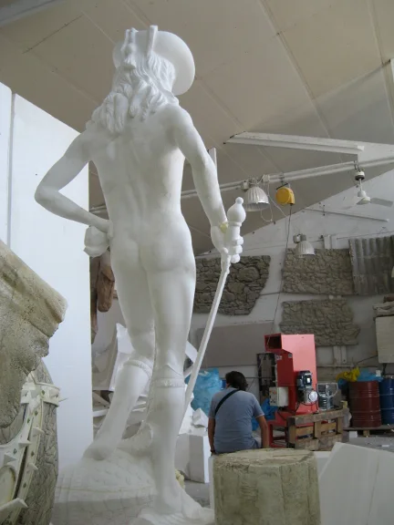 Statue of a classical hero sculpted at life size: nude male figure with Greek helmet, scepter in his right hand, heroic posture, rear view, being sculpted in the workshop hall among archived friezes and capitals
