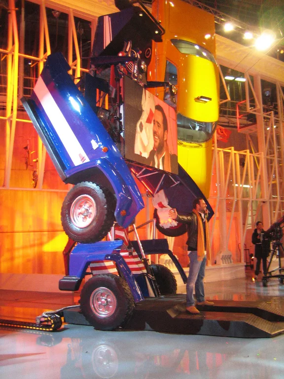 Vehicle-to-robot transformation phase: metallic blue doors open upward like wings, chassis wheels visible below, LED screen partially visible between the doors showing a close-up of the host; in the background a yellow industrial lattice structure