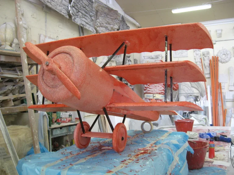 Red scenic biplane under construction in the workshop: overlapping wings, red fuselage, small wheels, World War I airplane profile, in the background an archive of classical statues, capitals, and friezes from the workshop