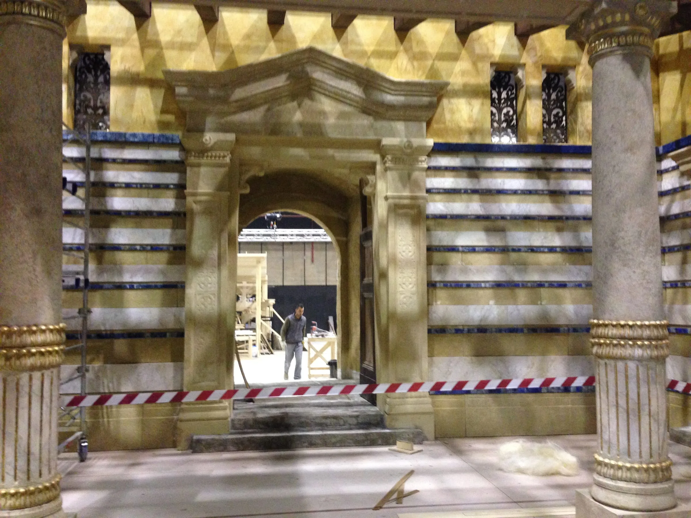 Arco monumentale del Tempio in pietra finta con timpano classico, fra pareti a fasce di marmo bianco con strisce blu di lapislazzulo. Cantiere con nastri rosso-bianchi e un operaio che cammina sullo sfondo