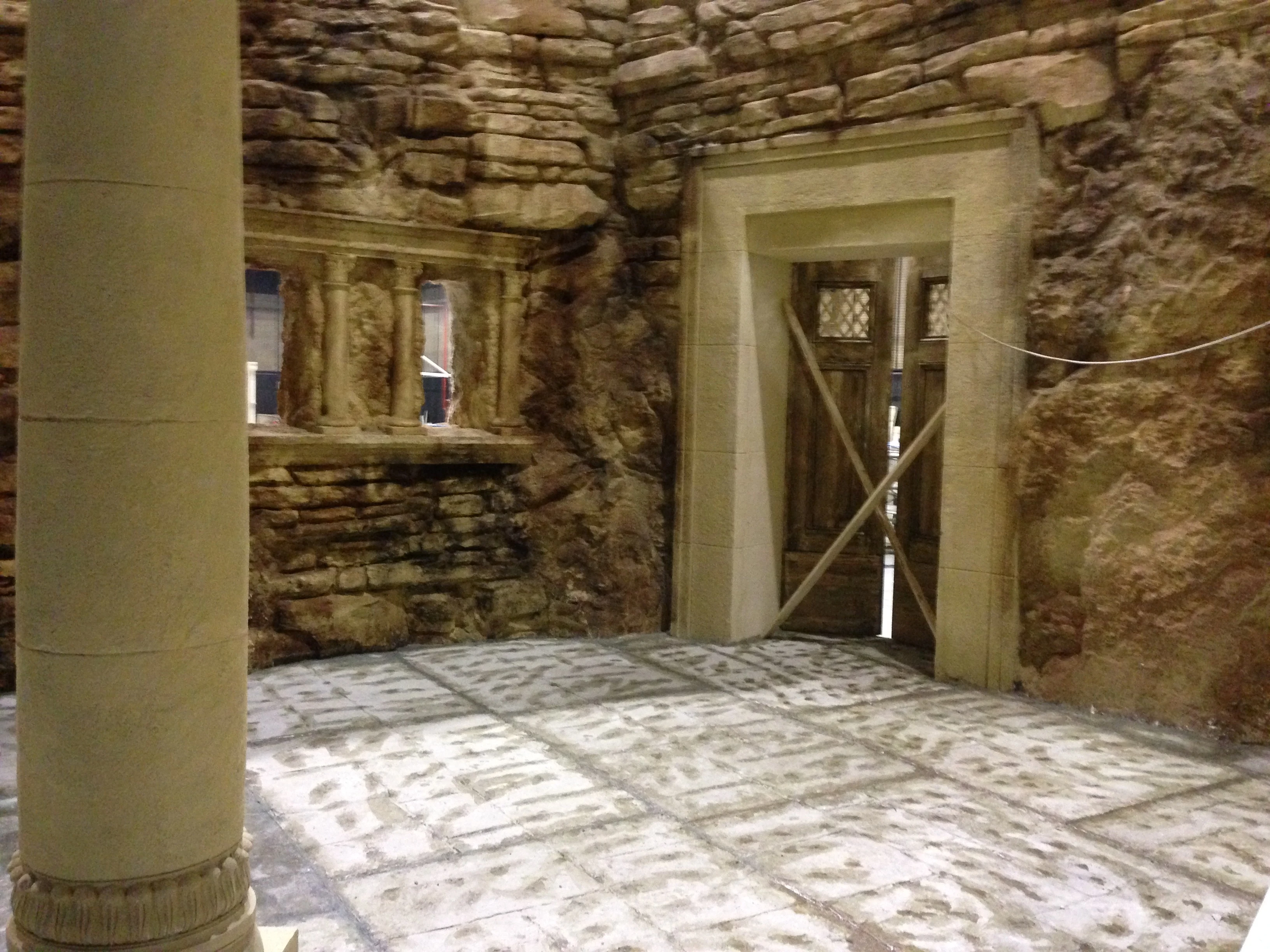 Set di una casa antica costruita in studio: pareti rocciose scolpite, una porta a doppio battente in legno, una nicchia con decorazioni, un mosaico geometrico a terra