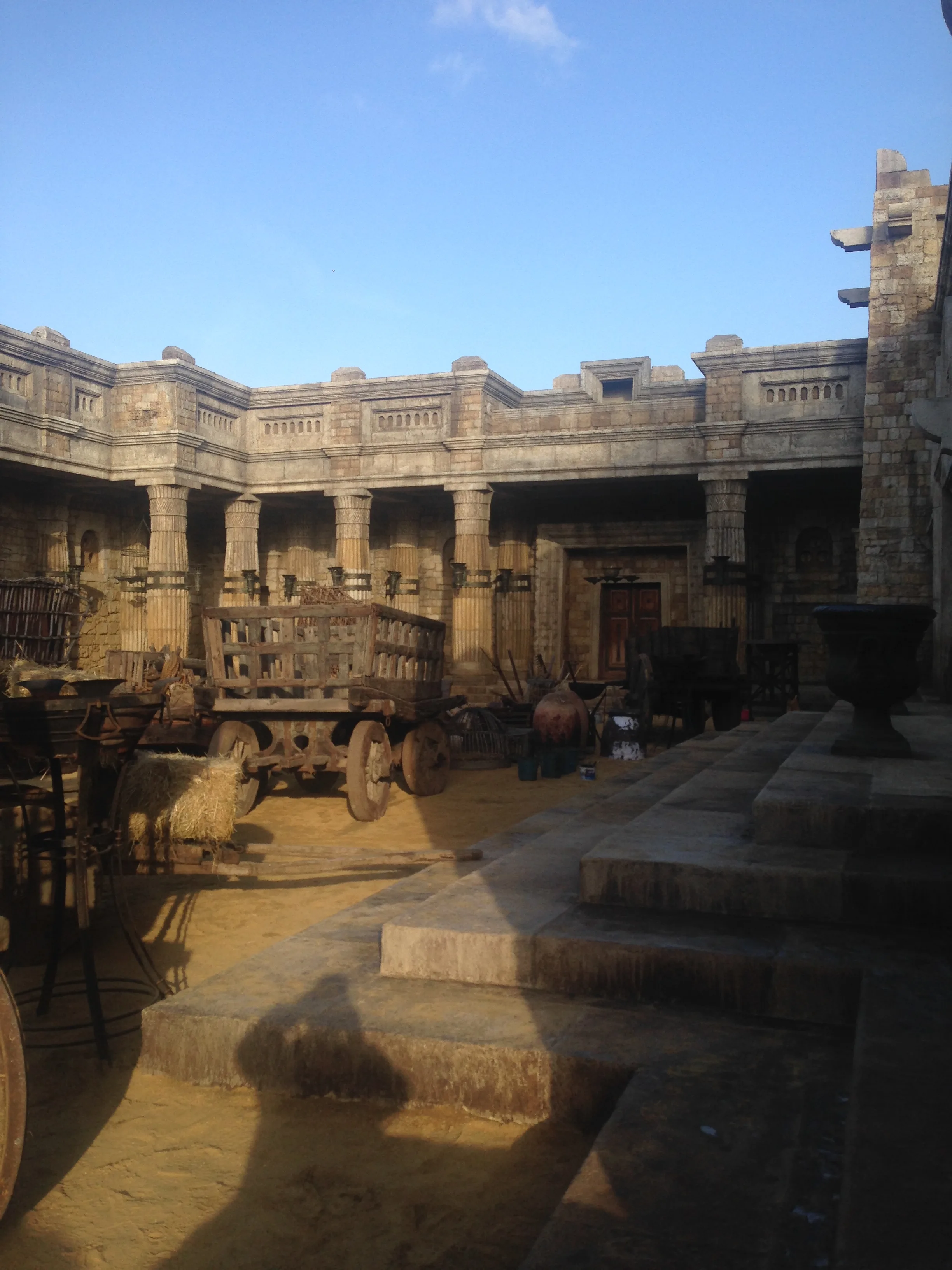 Vista grandangolare di un cortile esterno in stile romano-provinciale costruito a Cinecittà: mura merlate, portico colonnato, carro in legno carico di paglia con pecora viva accanto, scalini in pietra
