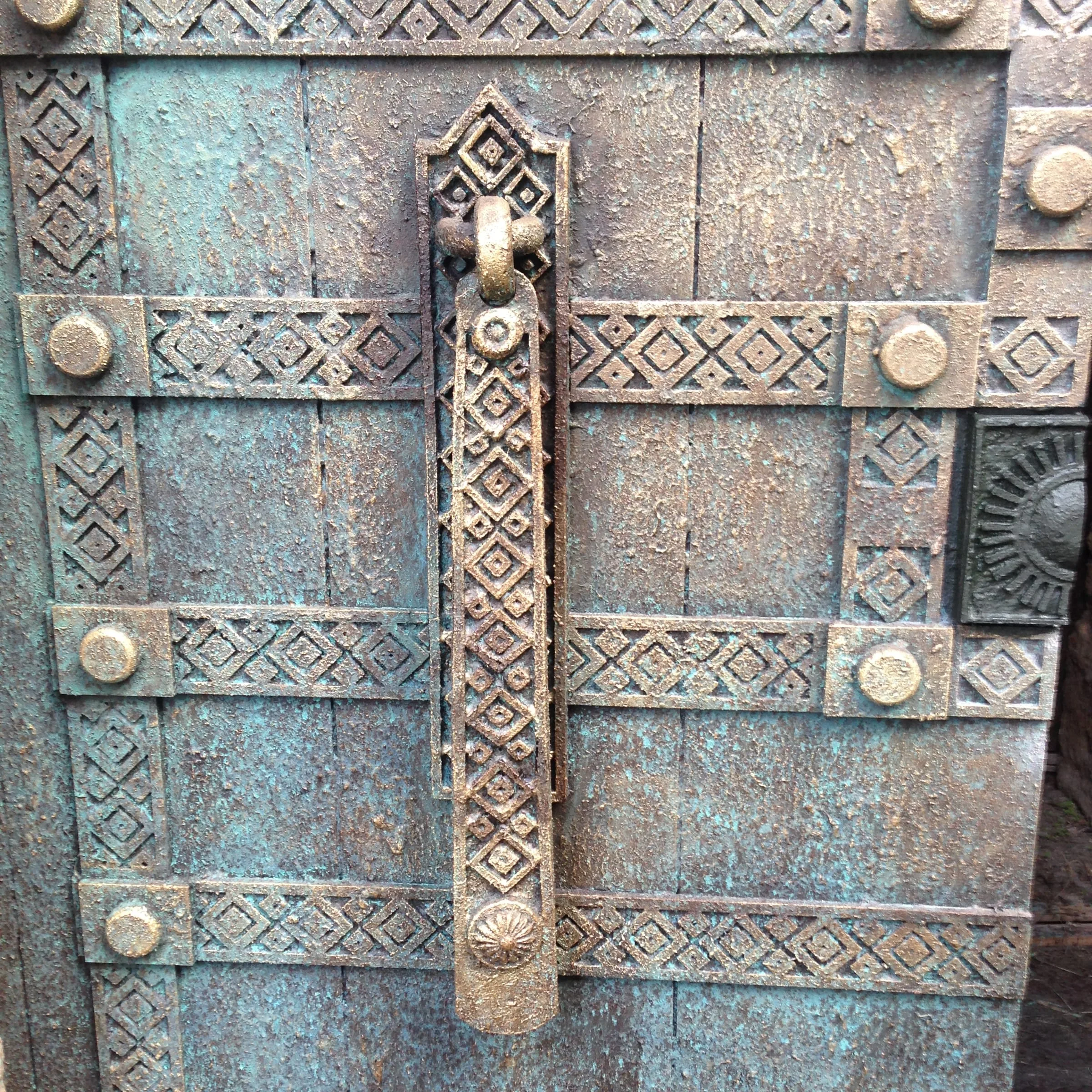 Primo piano di una porta in legno blu antico con dettagli geometrici a borchie metalliche: maniglione in bronzo verde rame, fasce ornamentali a losanghe