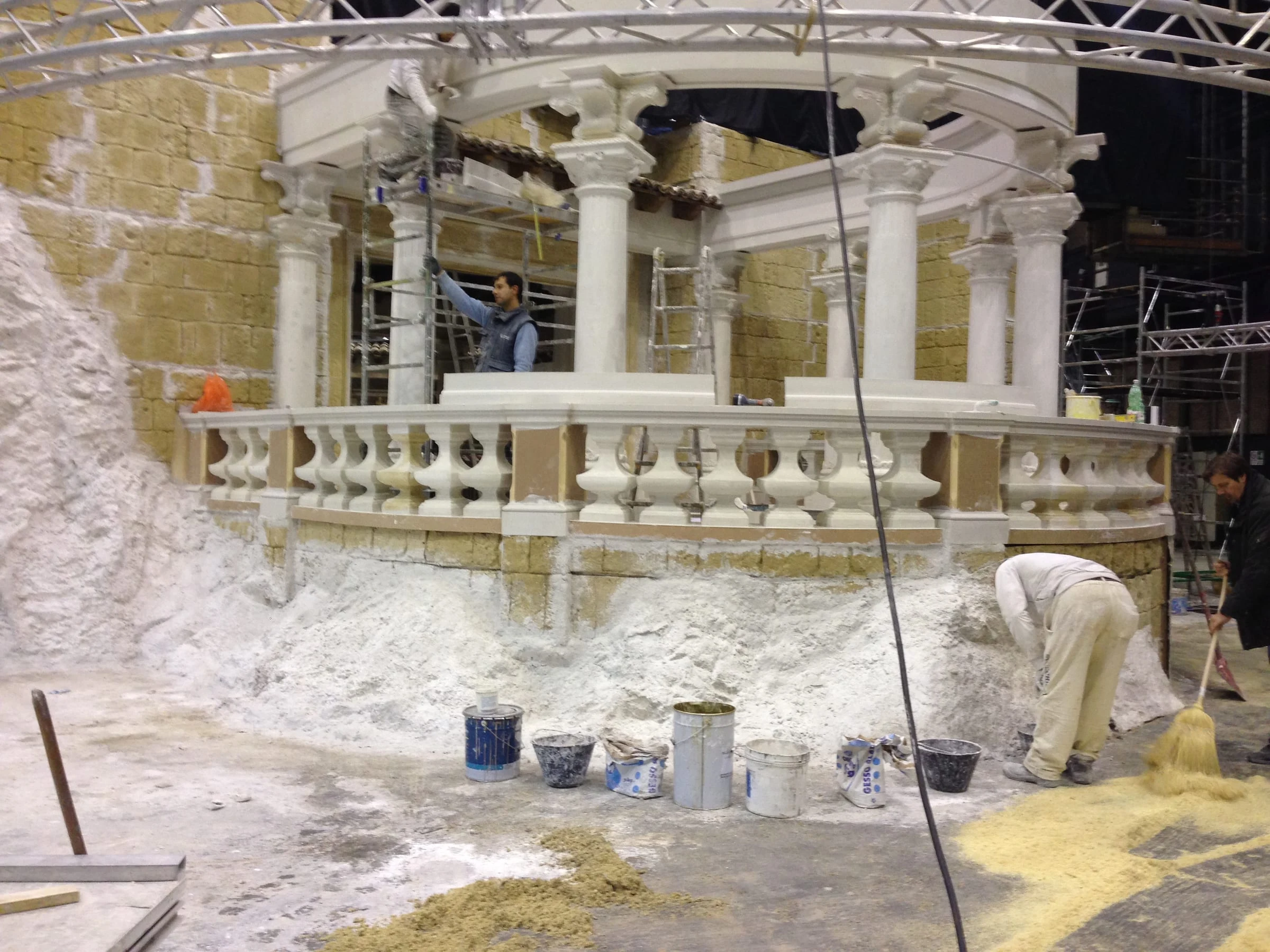 Set del Tempio in costruzione a Cinecittà: balaustra in pietra finta con colonnine, colonne classiche in marmo simulato, copertura a tegole, due operai in primo piano che intonacano la base con malta fresca