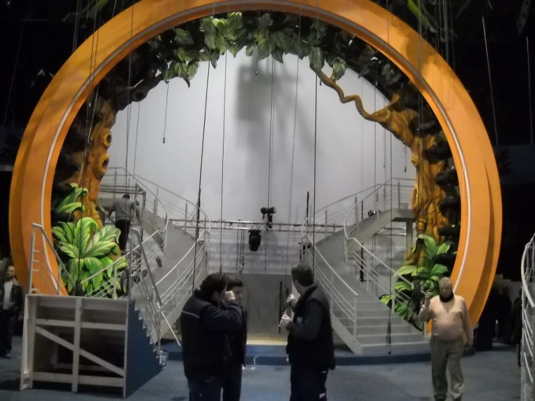 The Genodrome portal of Ciao Darwin during final construction: a large orange ring about three metres across at the centre of the TV studio, jungle tree trunk on the left of the ring sculpted in the round with green tropical leaves, twin staircases climbing the sides and converging at the centre, white-grey cyclorama in the background, technicians at work